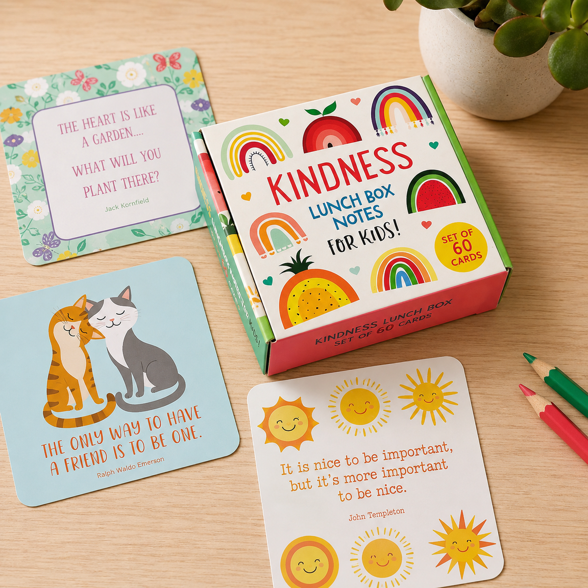 Kindness Lunch Box Notes for Kids set with a colorful box of 60 cards, shown alongside several sample cards on a light wood tabletop with a potted plant and colored pencils nearby.