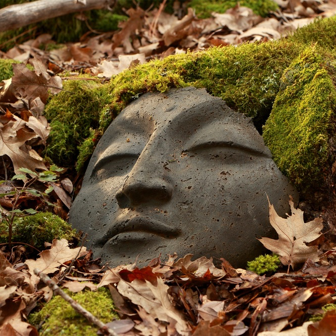 Stone face sculpture surrounded by moss and leaves in a natural setting