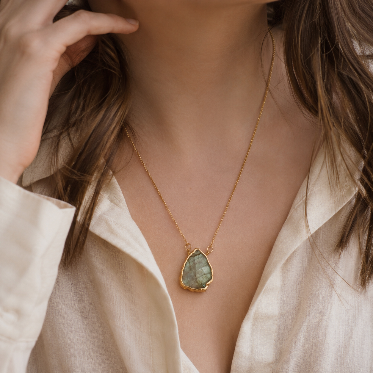 A close-up of a woman wearing a delicate gold chain necklace with a teardrop-shaped green stone pendant, paired with a cream-colored linen shirt.