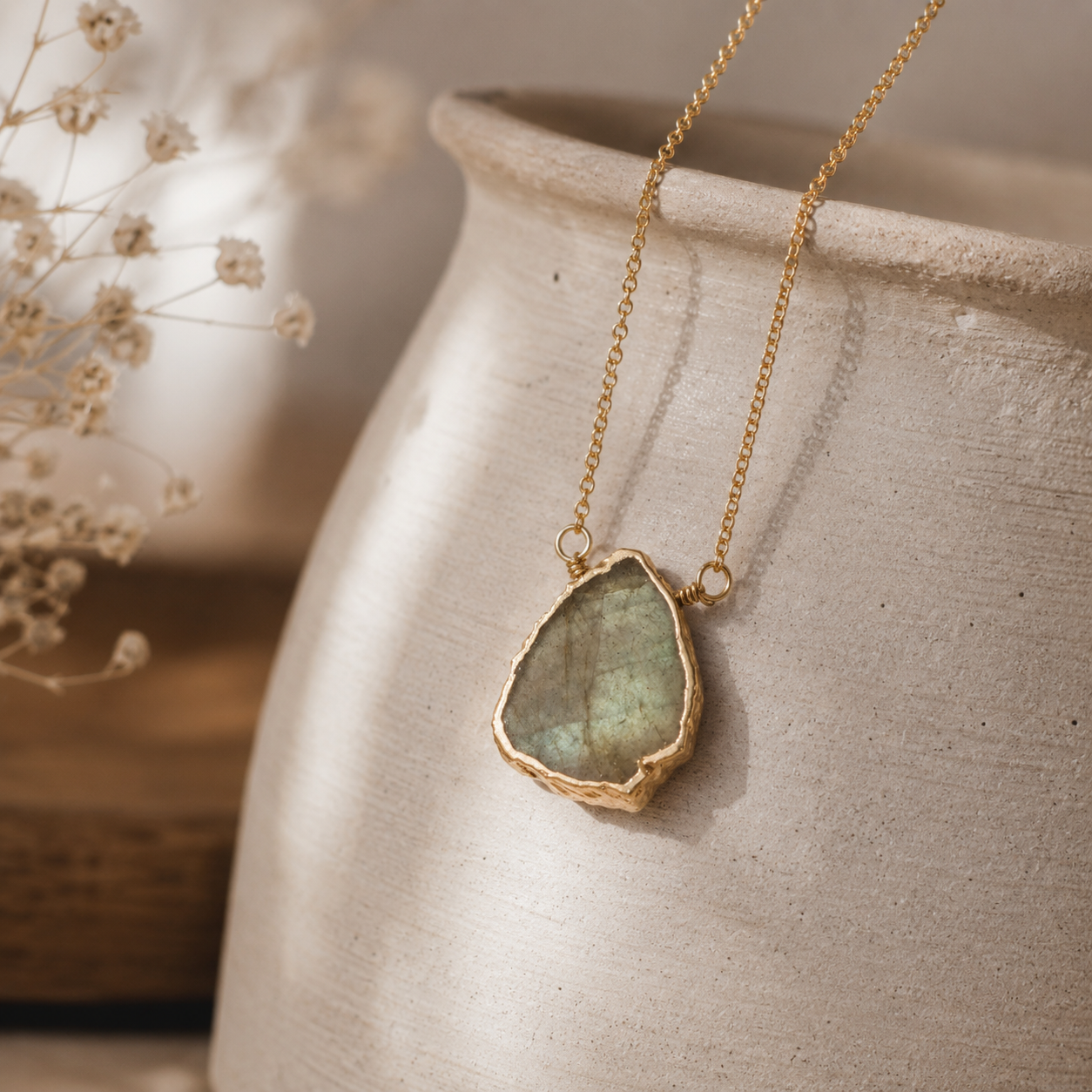A gold chain necklace with a raw, irregular green stone pendant, draped over the side of a textured beige ceramic vase.