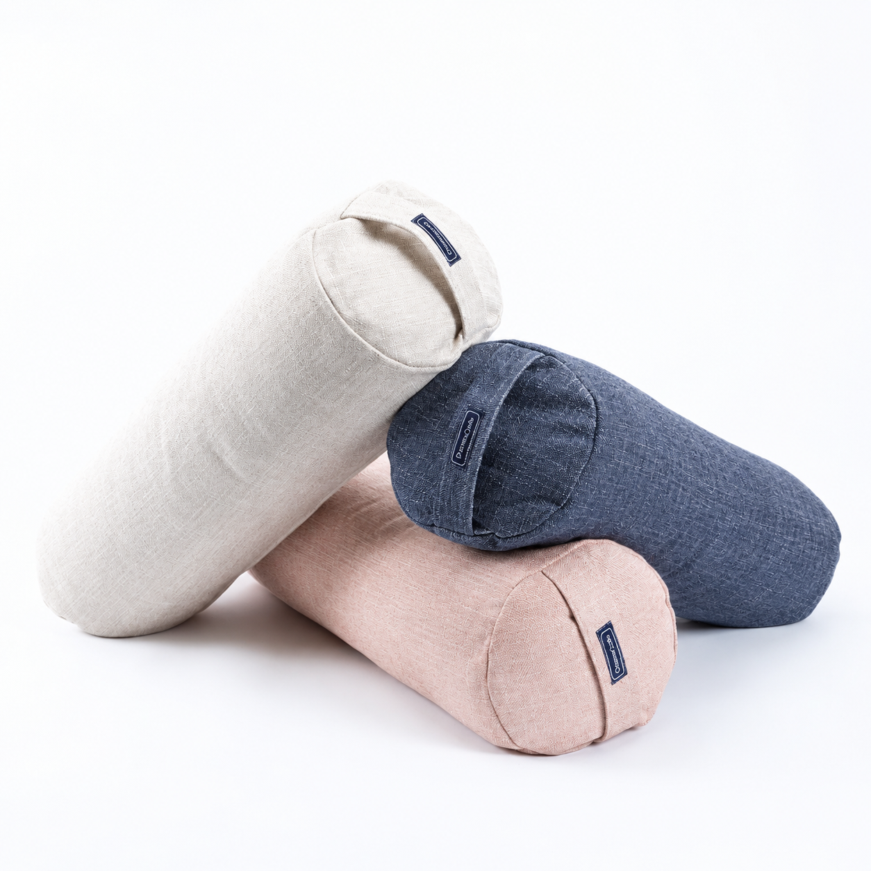 Three cylindrical bolster pillows in light beige, dusty pink, and slate blue, stacked together on a clean white background.