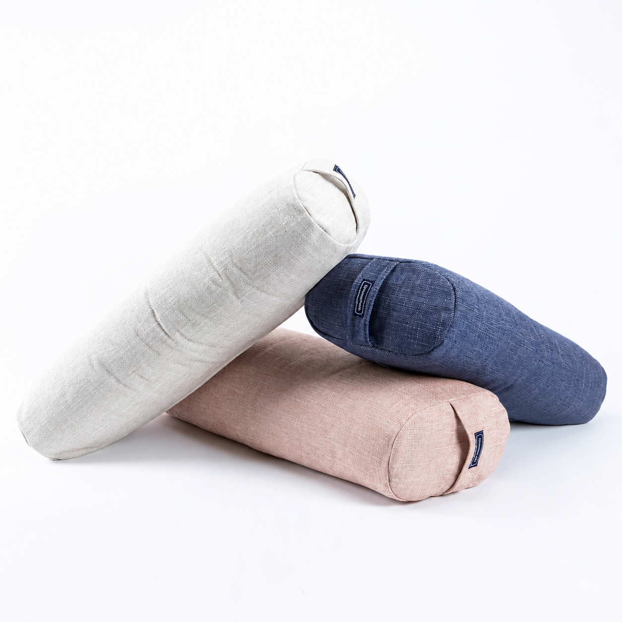 Three cylindrical yoga props or bolster pillows in light beige, dusty pink, and navy blue, stacked on a clean white background.