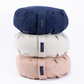 Three round, quilted cushion-like products stacked vertically on a white background, in navy blue, light beige, and blush pink. Each has a small fabric tag with the brand name 'Bloomsoft'.