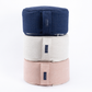 Three round meditation cushions stacked vertically on a white background, in navy blue, off-white, and light pink. Each cushion features a vertical strap with a small black brand label.