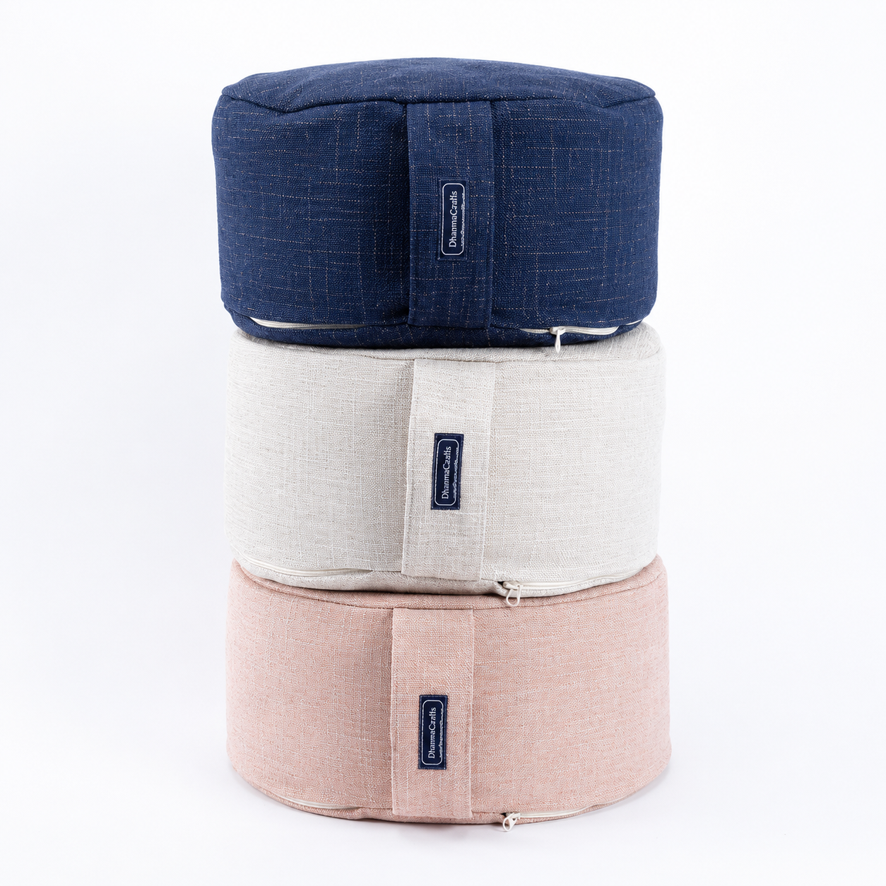 Three round meditation cushions stacked vertically on a white background, in navy blue, off-white, and light pink. Each cushion features a vertical strap with a small black brand label.