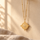 A gold pendant necklace with a circular mandala design hangs from a dark wooden peg on a textured beige wall, with soft sunlight and shadows cast across the scene.