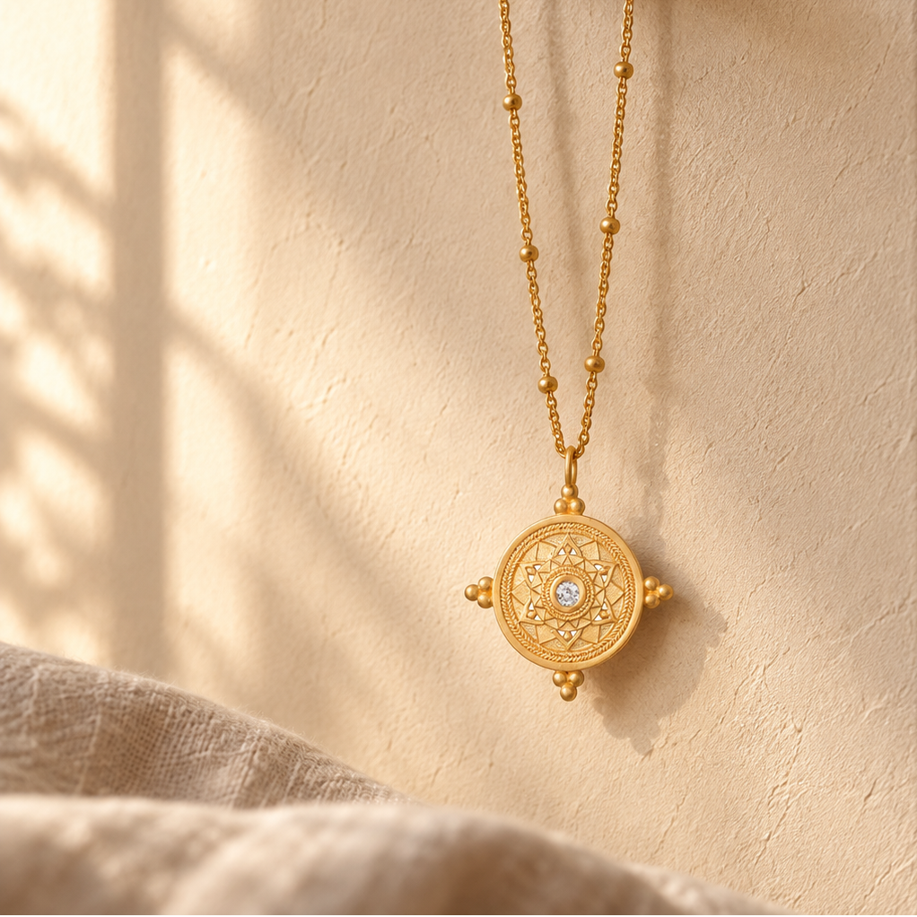 A gold pendant necklace with a circular mandala design hangs from a dark wooden peg on a textured beige wall, with soft sunlight and shadows cast across the scene.