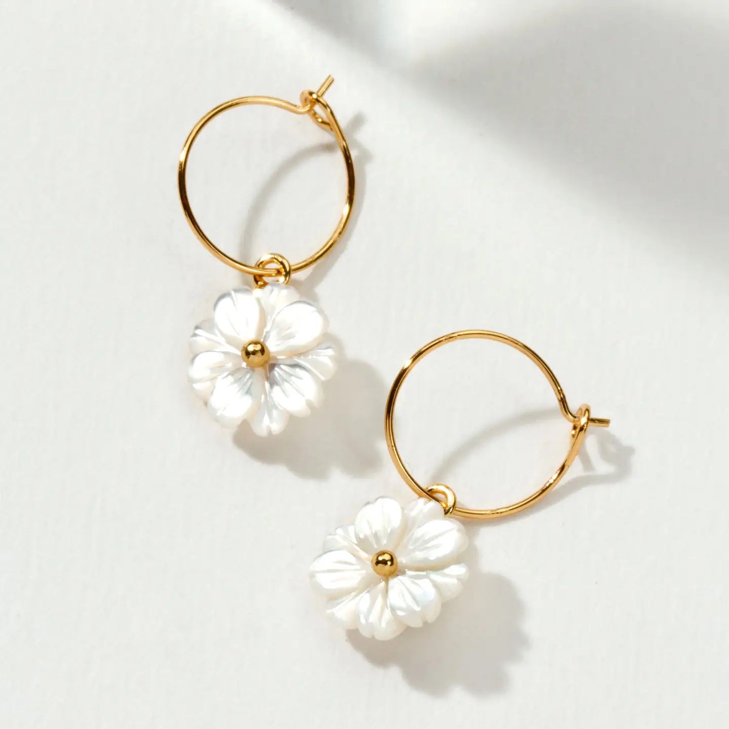 A pair of gold hoop earrings with dangling white flower charms, photographed on a clean white background with soft shadows.