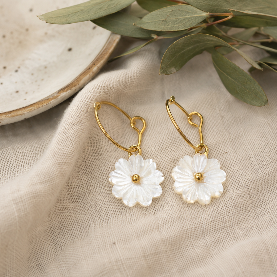 A pair of gold-tone daisy drop earrings with white pearl-like petals, displayed on a beige linen fabric next to a ceramic dish and green leaves.