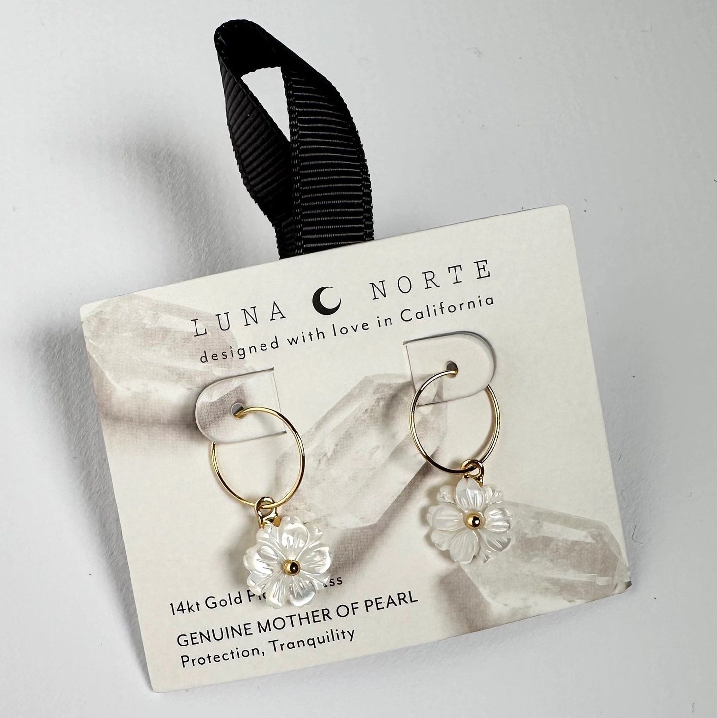 Luna Norte gold hoop earrings with white mother-of-pearl flower charms, presented on a white display card with a black ribbon loop.