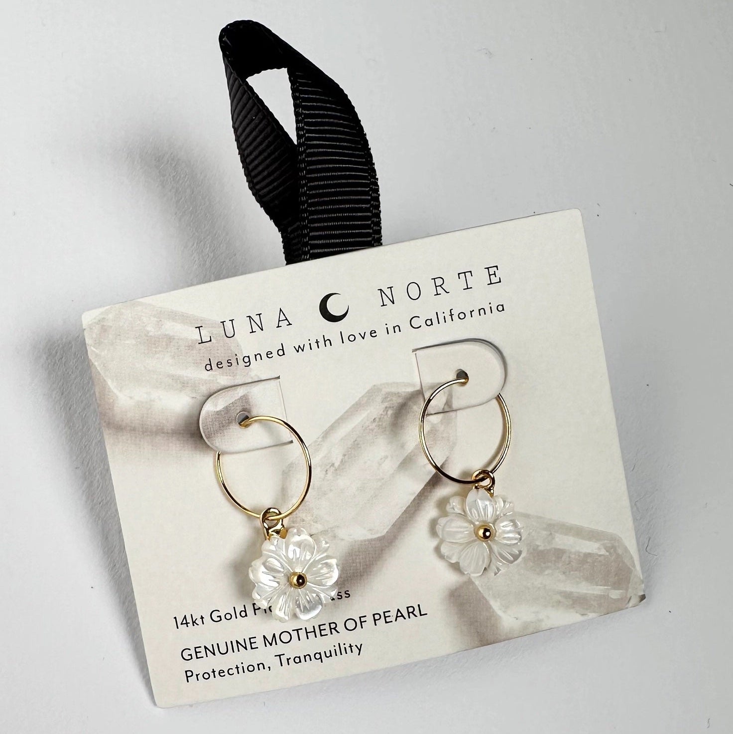 Luna Norte gold hoop earrings with white mother-of-pearl flower charms, presented on a white display card with a black ribbon loop.