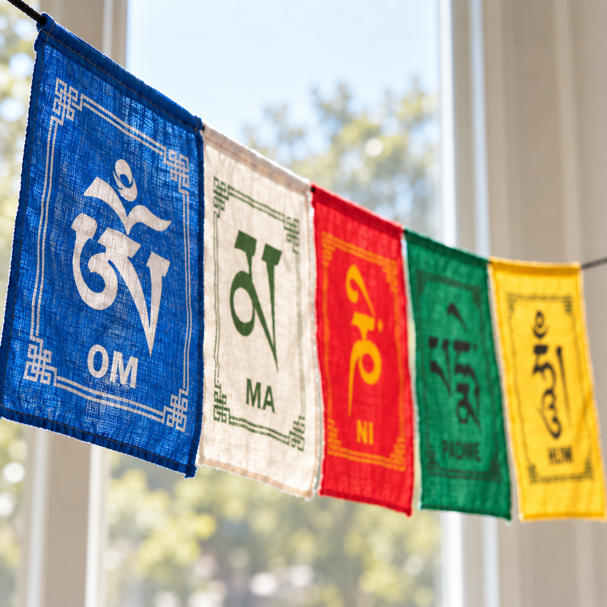 Five colorful fabric prayer flags hanging in a row, featuring white, green, red, green, and yellow panels with spiritual symbols and text, photographed in front of a softly blurred window with trees outside.