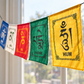 Four colorful Tibetan prayer flags hanging in a row outdoors near a window. The flags are blue, white, red, and yellow, printed with black Tibetan script and traditional border designs.