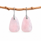 A pair of teardrop-shaped pink stone drop earrings with silver hooks, hanging from a natural brown branch against a clean white background.