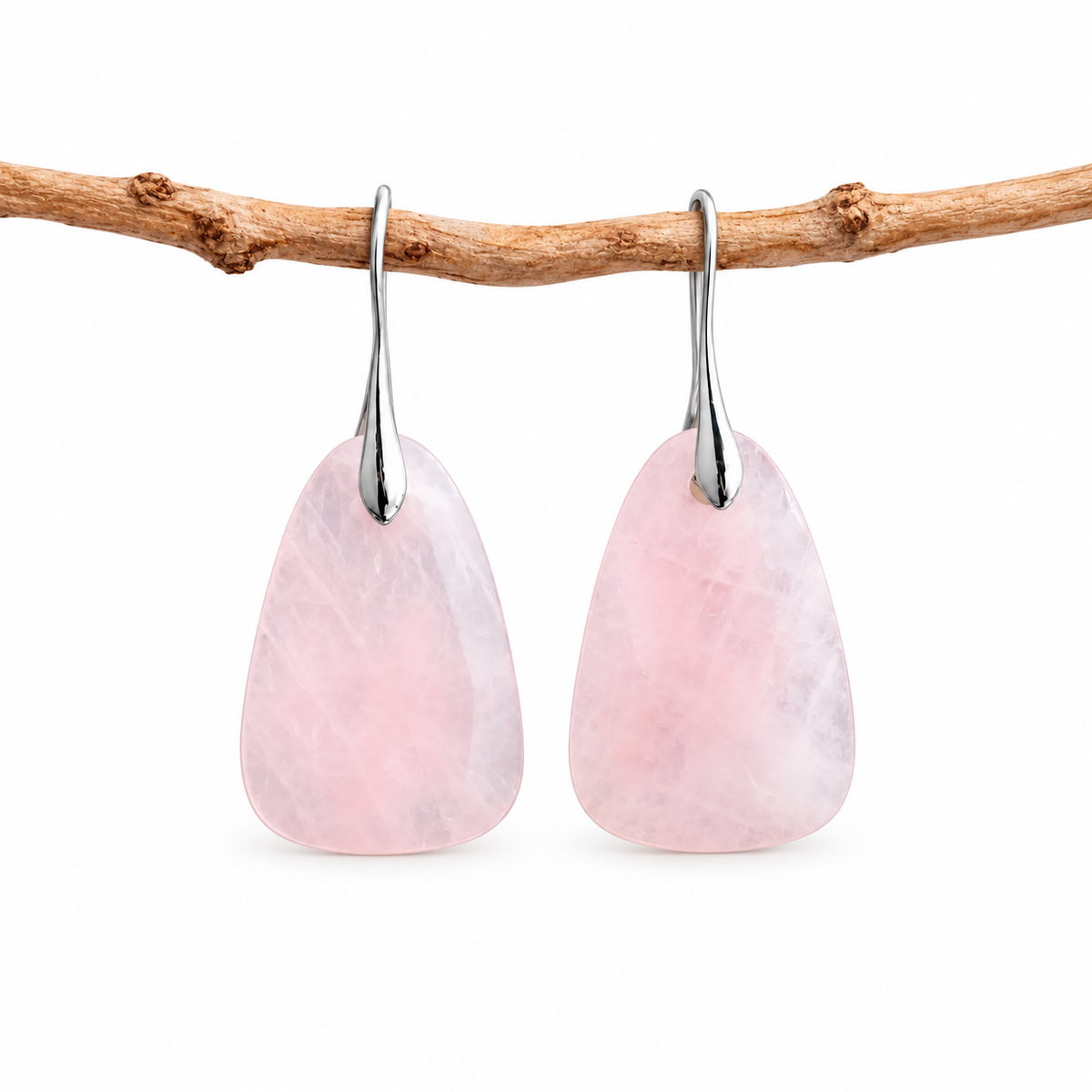 A pair of teardrop-shaped pink stone drop earrings with silver hooks, hanging from a natural brown branch against a clean white background.