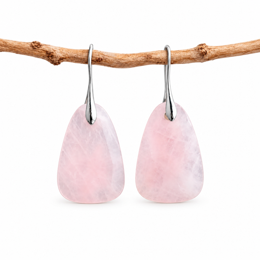 A pair of teardrop-shaped pink stone drop earrings with silver hooks, hanging from a natural brown branch against a clean white background.