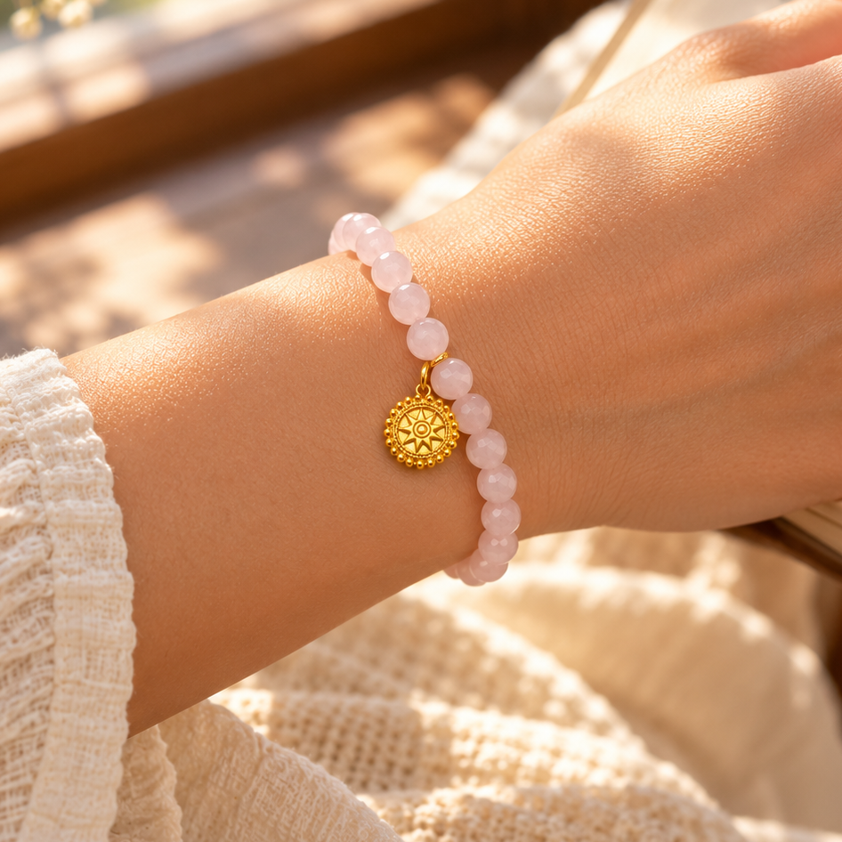 A person's wrist wearing a light pink beaded bracelet with a gold sun pendant, resting on an open book in a warm, sunlit indoor setting.