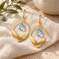A pair of gold drop earrings with a teardrop-shaped openwork frame and a small light blue teardrop gemstone, displayed on a textured cream linen cloth with decorative green leaves and a ceramic bowl in the background.