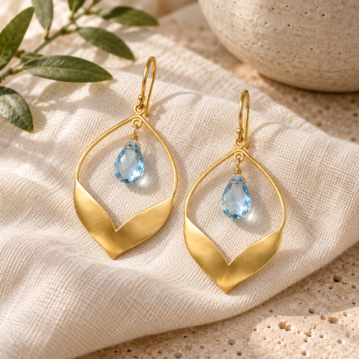 A pair of gold drop earrings with a teardrop-shaped openwork frame and a small light blue teardrop gemstone, displayed on a textured cream linen cloth with decorative green leaves and a ceramic bowl in the background.