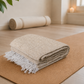 Folded beige blanket with white fringe on a yoga mat in a serene room setting.