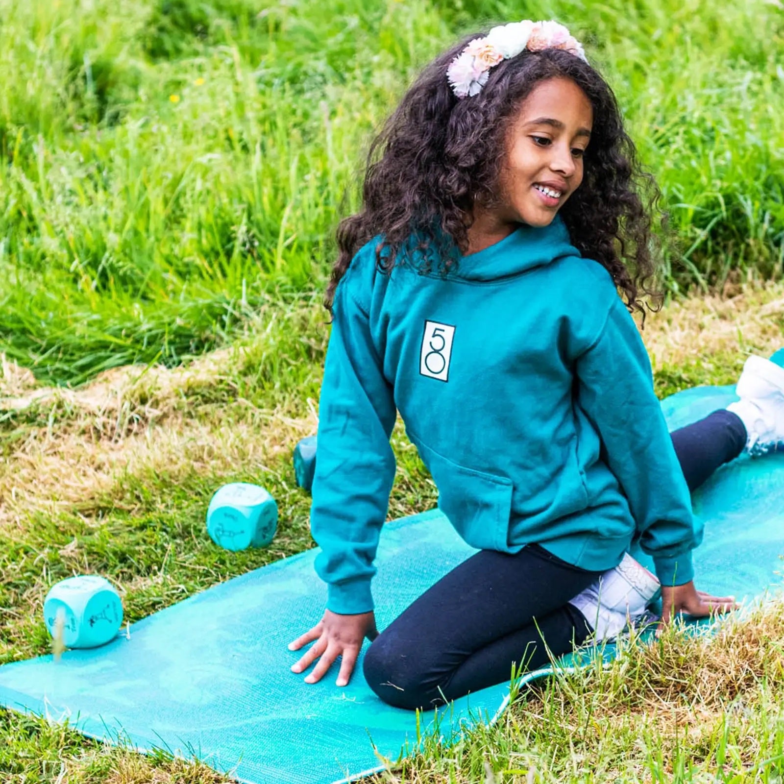 Child in a teal hoodie sitting on a yoga mat outdoors