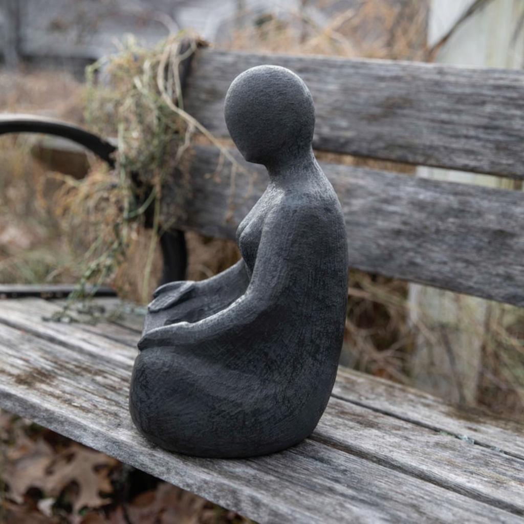 Gray stone sculpture of a seated figure on a wooden bench with a natural background