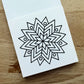 A white square card or notebook page with a black geometric mandala-style line drawing, photographed on a light wood surface.