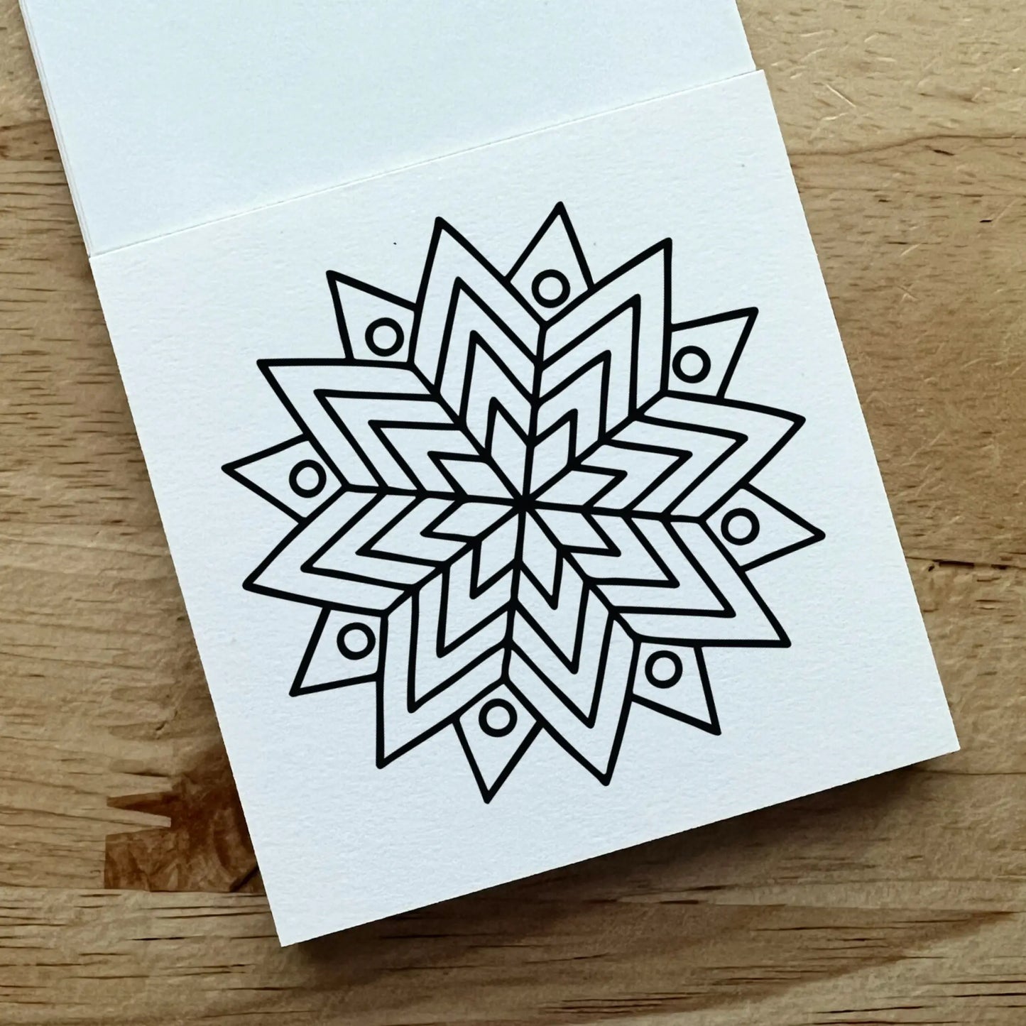 A white square card or notebook page with a black geometric mandala-style line drawing, photographed on a light wood surface.