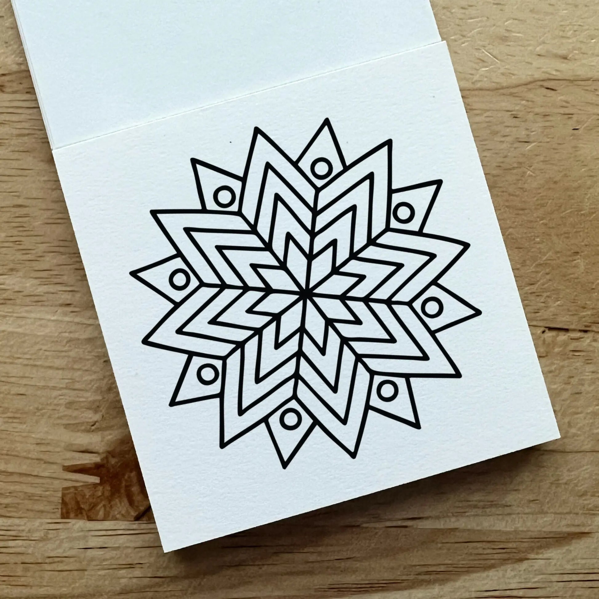 A white square card or notebook page with a black geometric mandala-style line drawing, photographed on a light wood surface.