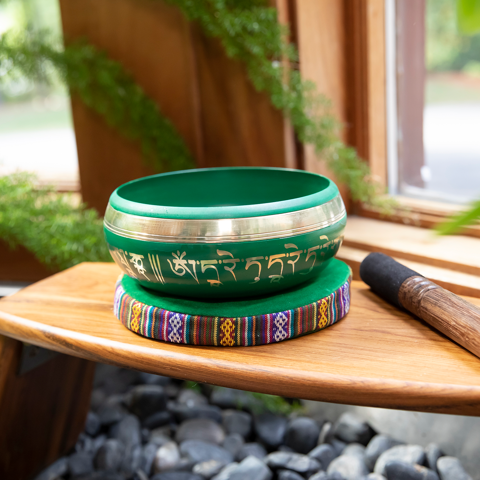 The Green Tara Singing Bowl rests atop a cushion and wooden meditation stool, mallet at its side. Plants and rocks can be seen under and around the subject.