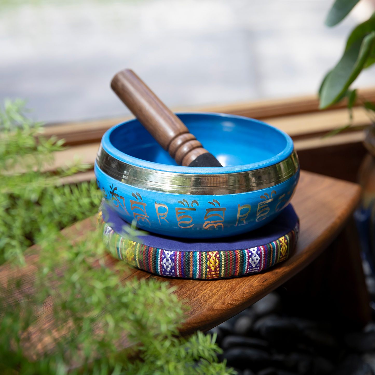 The Medicine Buddha Singing Bowl rests atop a cushion and wooden meditation stool, mallet inside, while plants frame the subject.